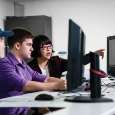 Students focused on computers in a classroom with their professor pointing something out on the screen.