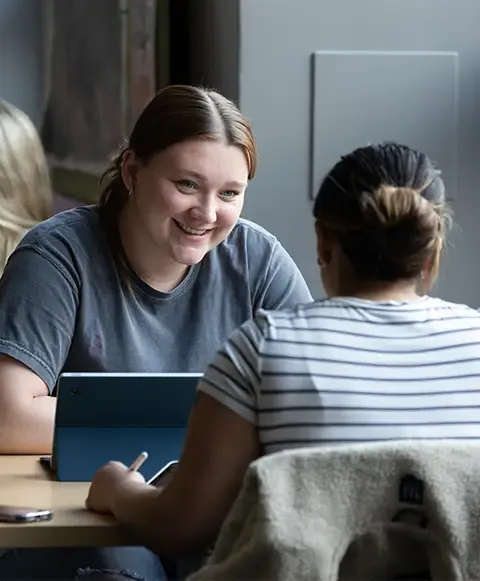 Two women sitting at a table with laptops, engaged in discussion.